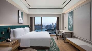 A modern hotel room with a large bed, a desk, and a view of a cityscape through large windows.