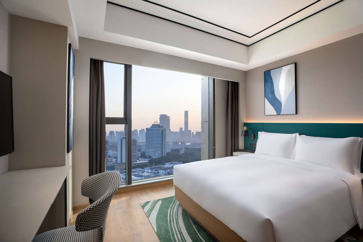 A modern hotel room with a large bed, a view of the cityscape, a desk, and a chair.