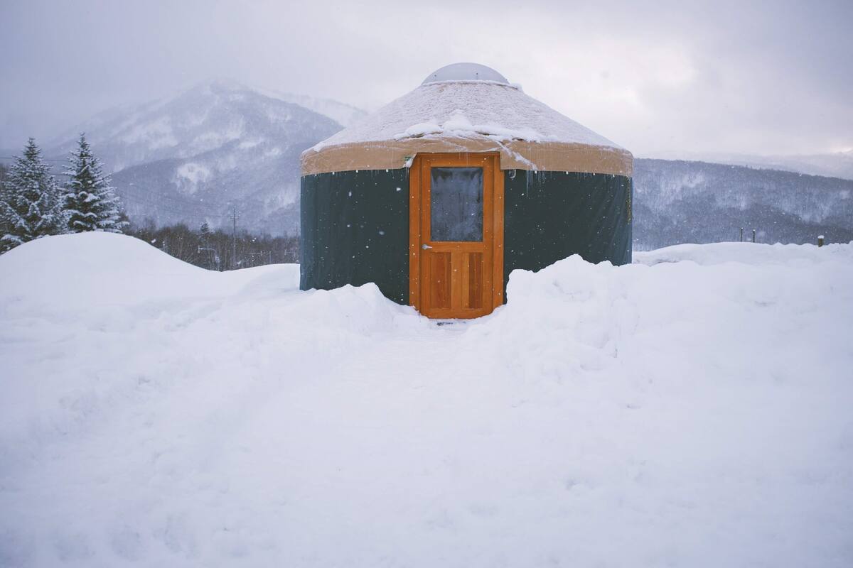 Una yurta cubierta de nieve con una puerta de madera, rodeada de nieve y montañas al fondo.
