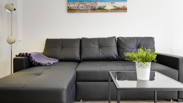 Ein modernes Wohnzimmer mit einer schwarzen Ledersofa, einem Glastisch und einer Zimmerpflanze. Ein Strandbild hängt an der Wand.