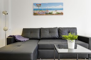 A modern living room with a black leather sofa, a glass coffee table, and a potted plant. A beach-themed painting is on the wall.