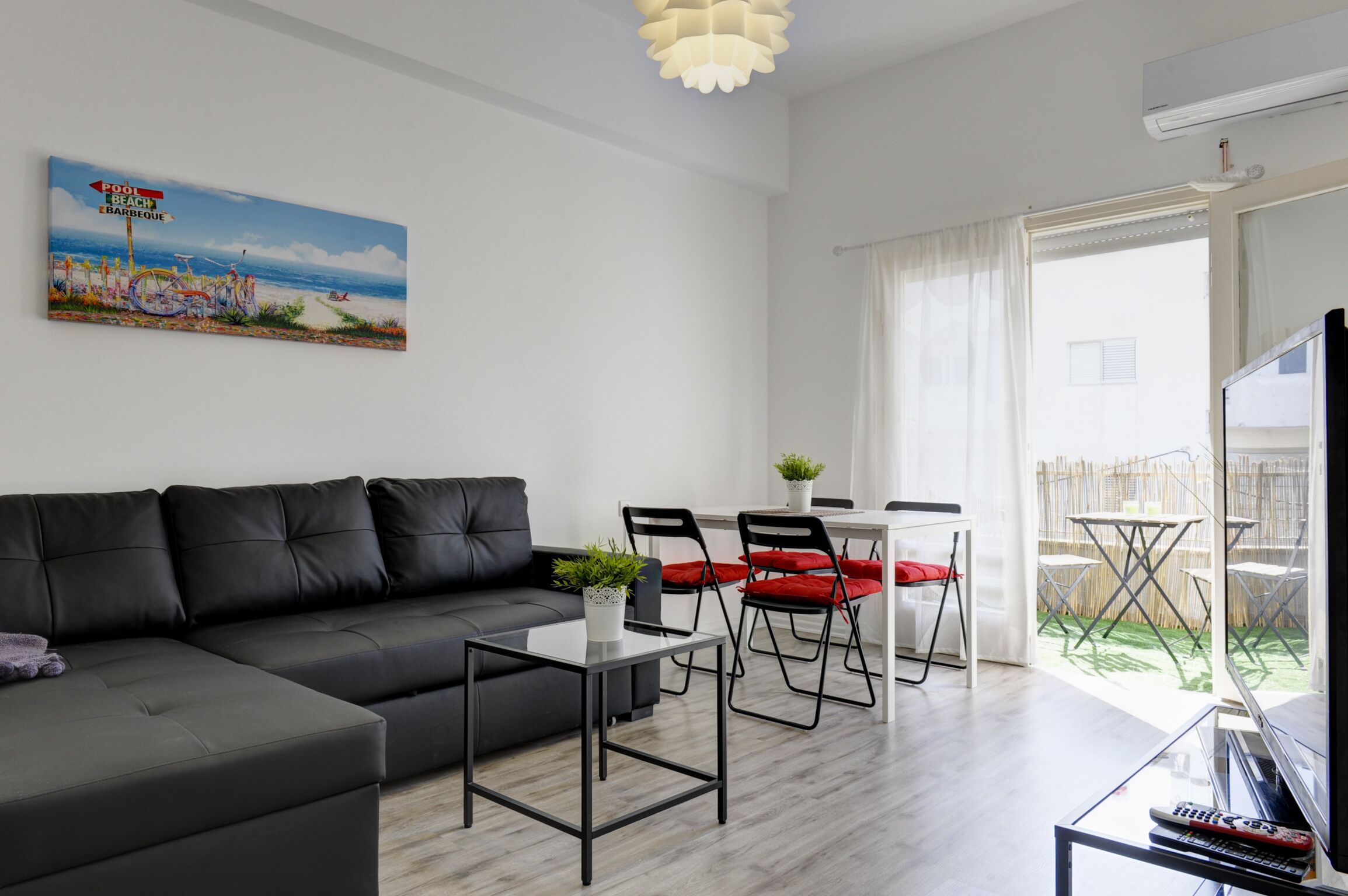 Un salon moderne avec un canapé noir, une table basse en verre et un coin repas avec des chaises rouges. Un tableau sur le thème de la plage est accroché au mur, et on aperçoit un balcon avec des meubles d’extérieur.