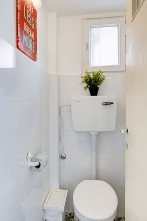 Une petite salle de bain blanche, avec des toilettes, un petit lavabo et une fenêtre située au-dessus des toilettes.