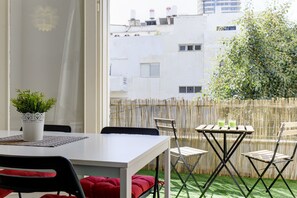 Un balcon avec une table et des chaises blanches, un pot de fleurs et une vue sur des immeubles et des arbres.