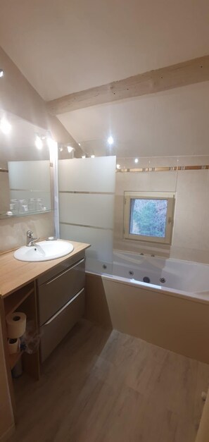 Une salle de bain moderne avec un meuble-lavabo en bois, un grand miroir, une baignoire et une fenêtre située au-dessus de la baignoire.
