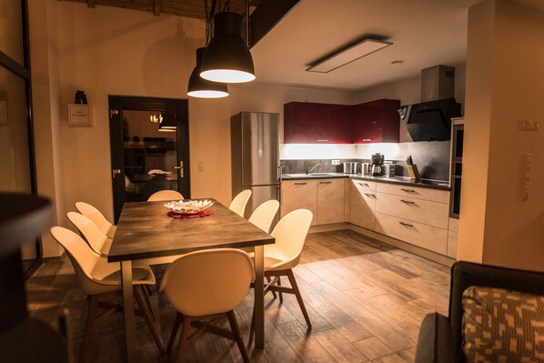 A modern kitchen with white cabinets, a stainless steel refrigerator, and a dining area with a wooden table and white chairs. - Chalet100 with private beach, sauna, fireplace and gallery on Lake Mühlfeld (Löbnitz)