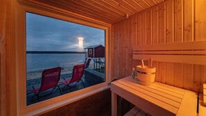 Une cabane en bois dotée d’une grande fenêtre donnant sur un plan d’eau, deux chaises rouges et un banc en bois.