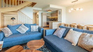 A modern living room with a staircase, blue sofas, wooden coffee table, and a kitchen in the background.