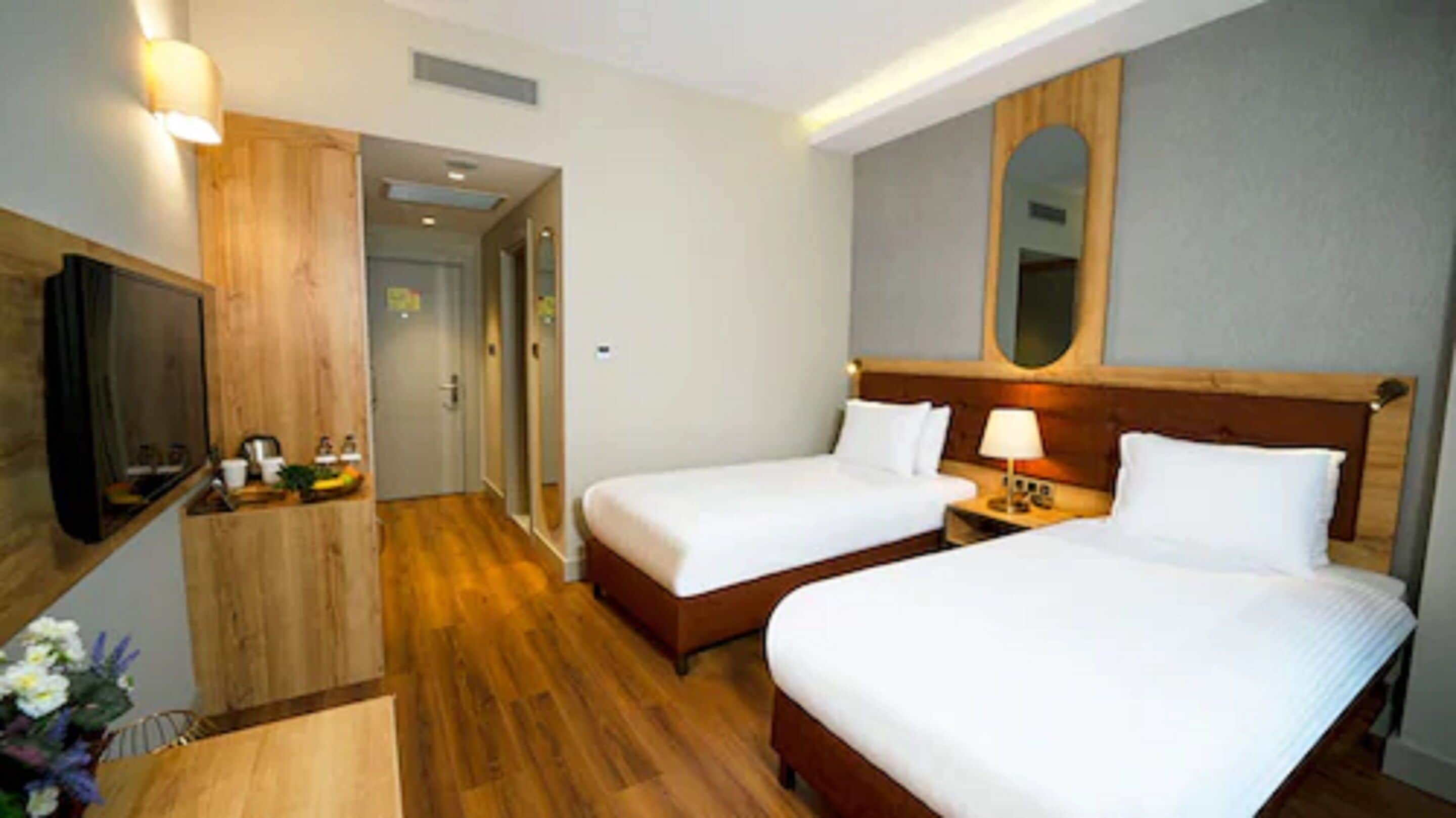 A hotel room with two beds, wooden flooring, a TV, and a small table with a lamp.