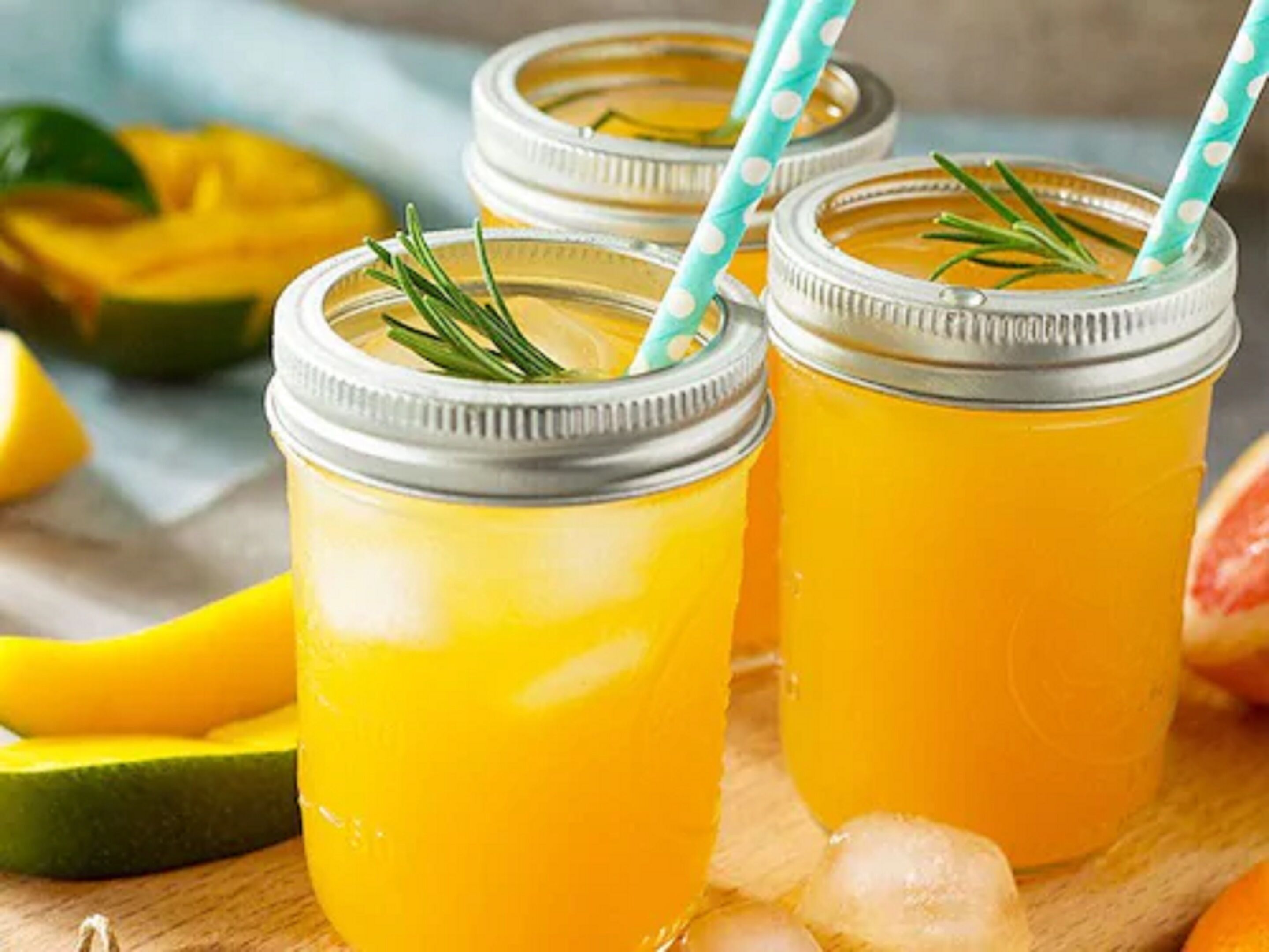 Three mason jars with a yellow liquid, possibly a drink, garnished with rosemary, and ice cubes on a wooden surface.