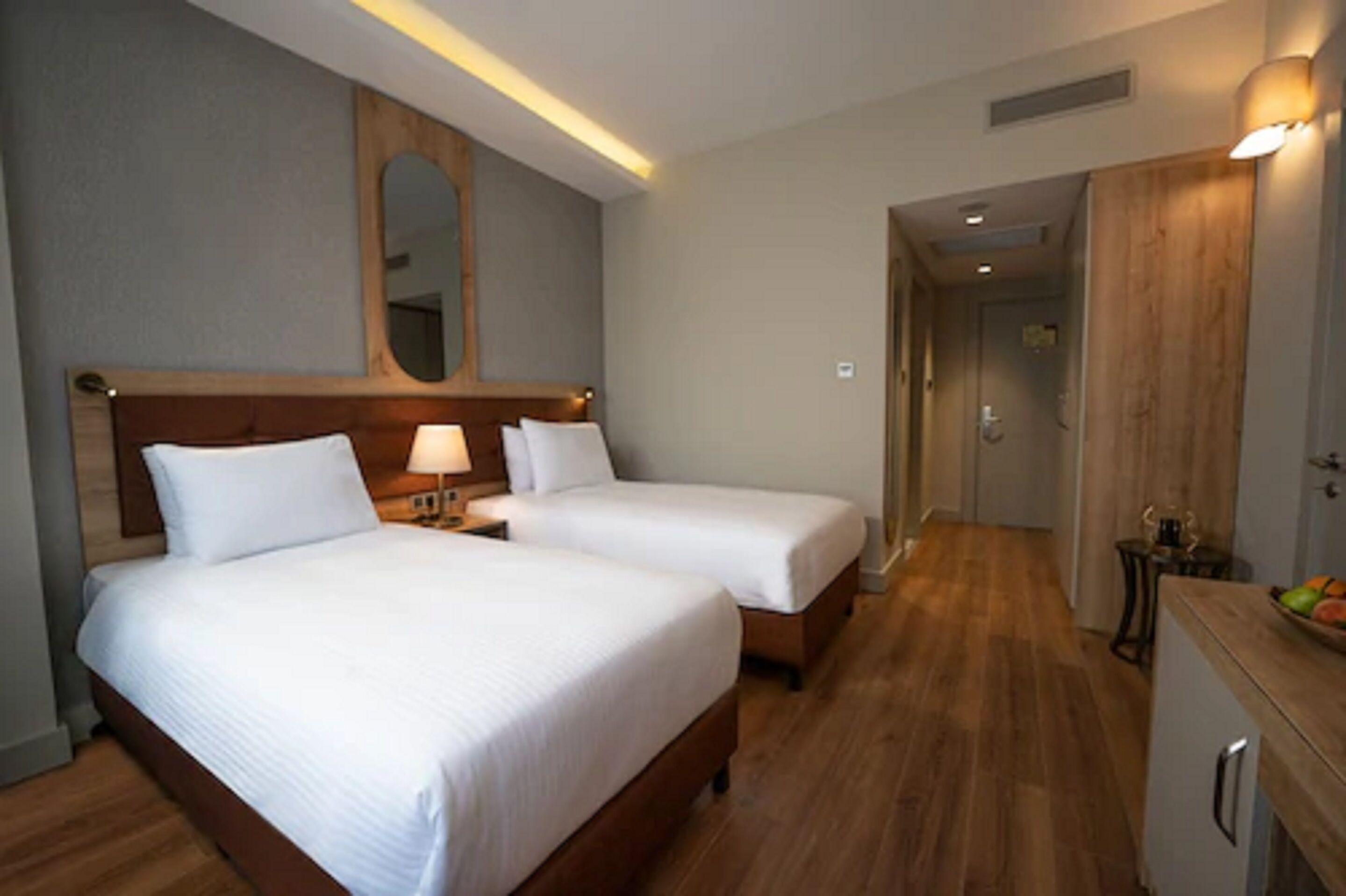 A hotel room with two single beds, a wooden headboard, a mirror, and a small kitchenette area.
