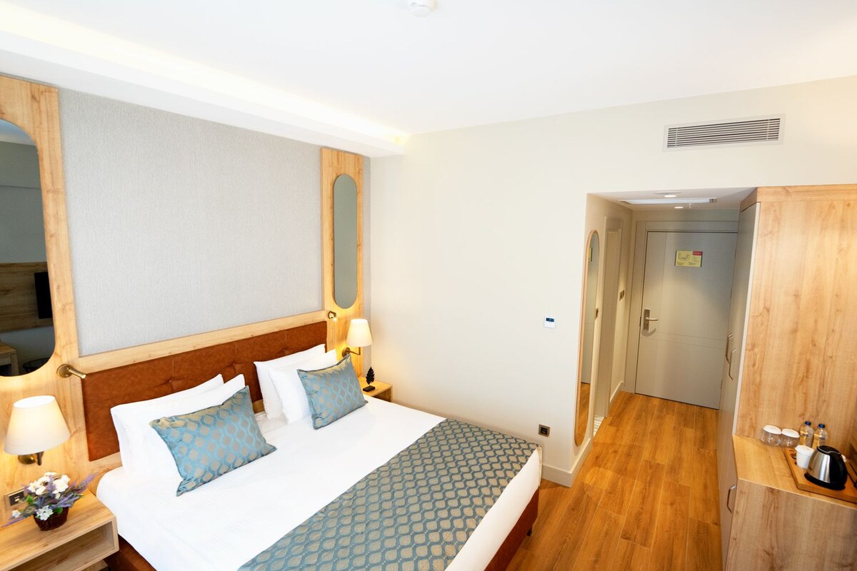 A hotel room with a wooden headboard, a large bed with patterned bedding, a bedside table with a lamp, and a wooden wardrobe.