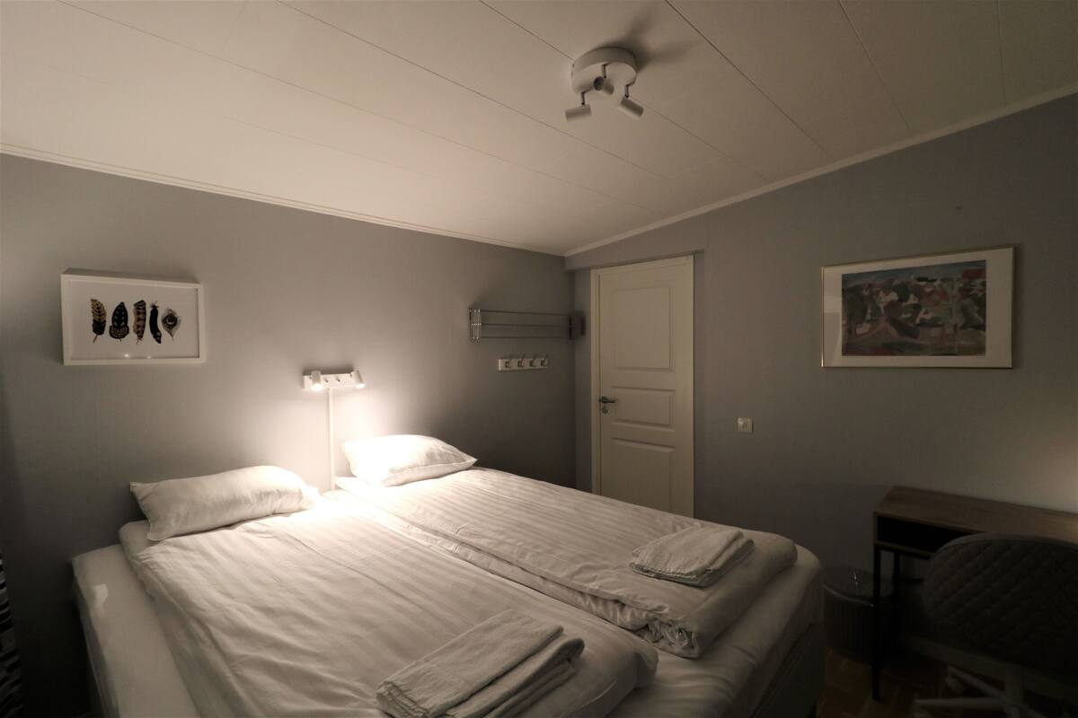Un dormitorio con dos camas, una mesita de noche, una lámpara, un cuadro enmarcado y una puerta.