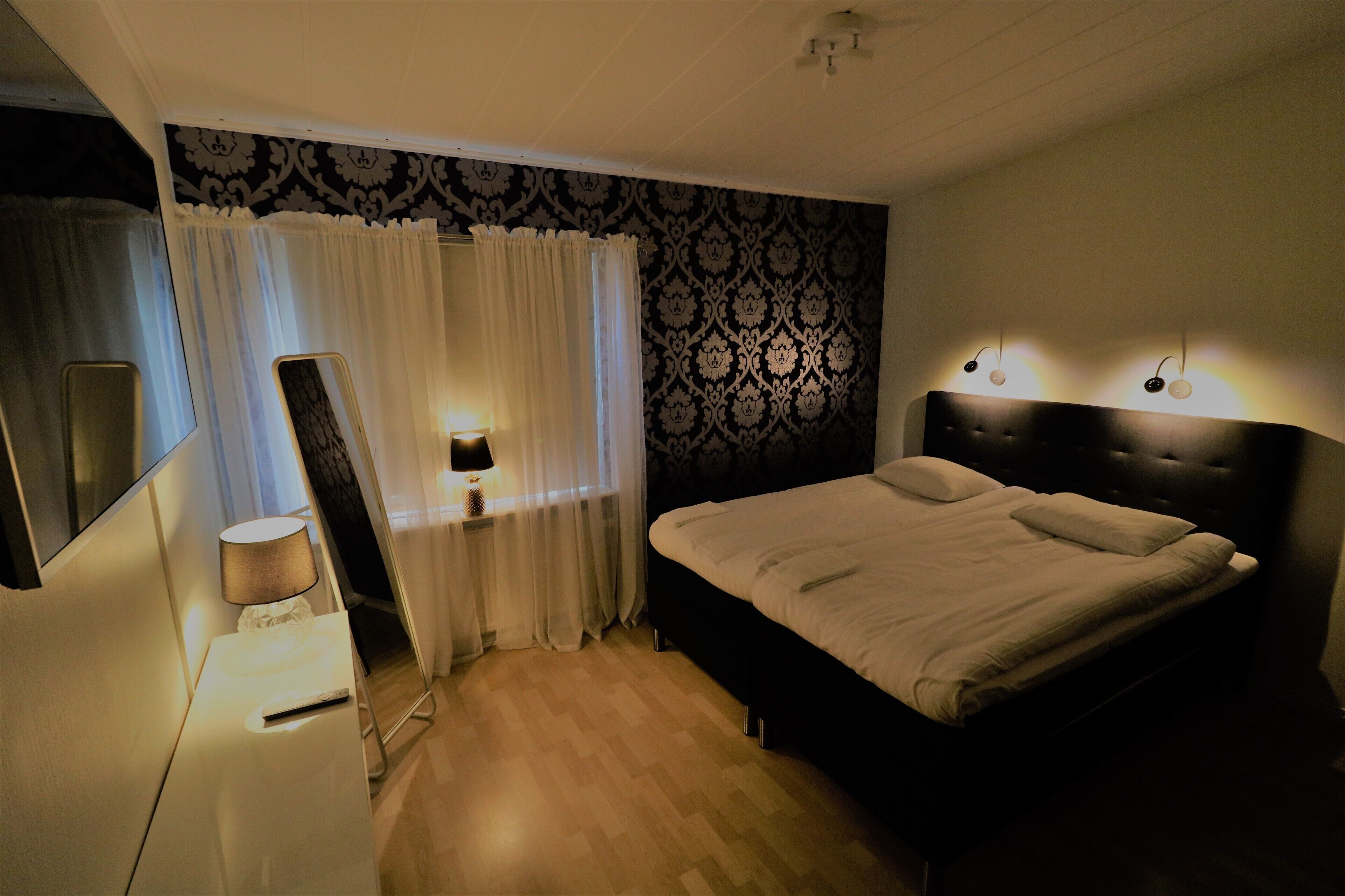 Een slaapkamer met een bed, nachtkastje met lamp, spiegel en een kamer met een patroon.