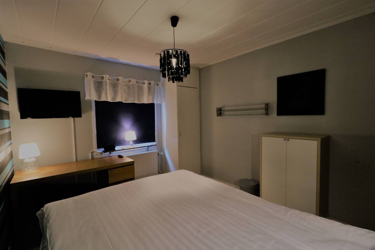 Habitación de hotel con cama, escritorio con lámpara, televisor, armario y ventana con cortina.
