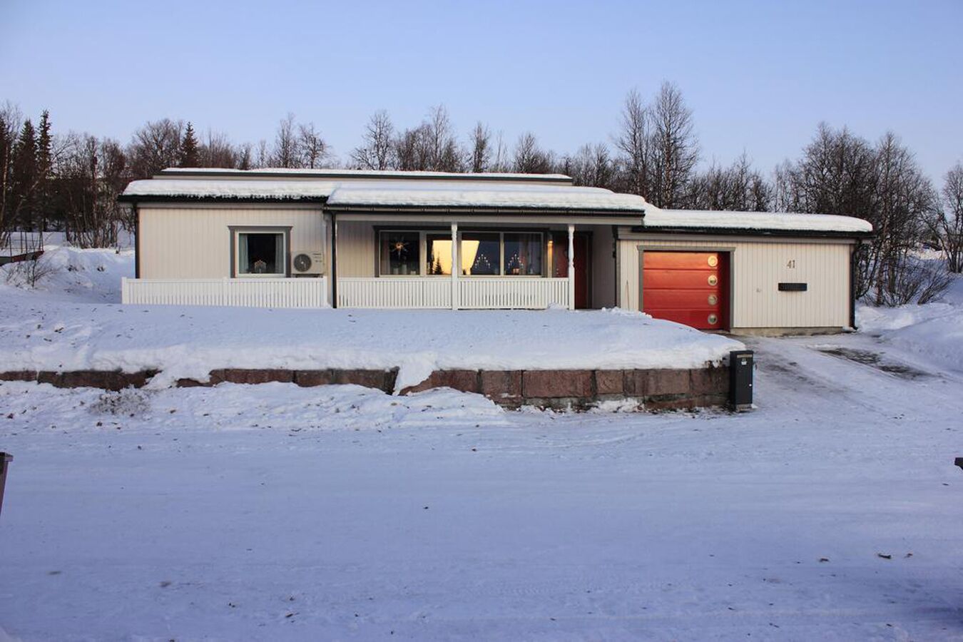Un bâtiment d’un seul étage, avec une porte de garage rouge, un petit porche et un toit recouvert de neige.