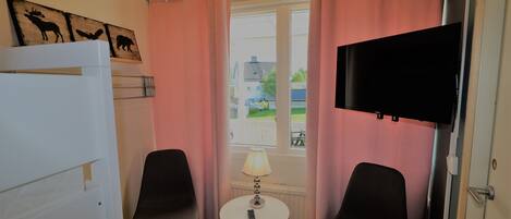 Une pièce comprenant un lit, deux chaises, une petite table avec une lampe, un téléviseur fixé au mur et une fenêtre avec des rideaux.