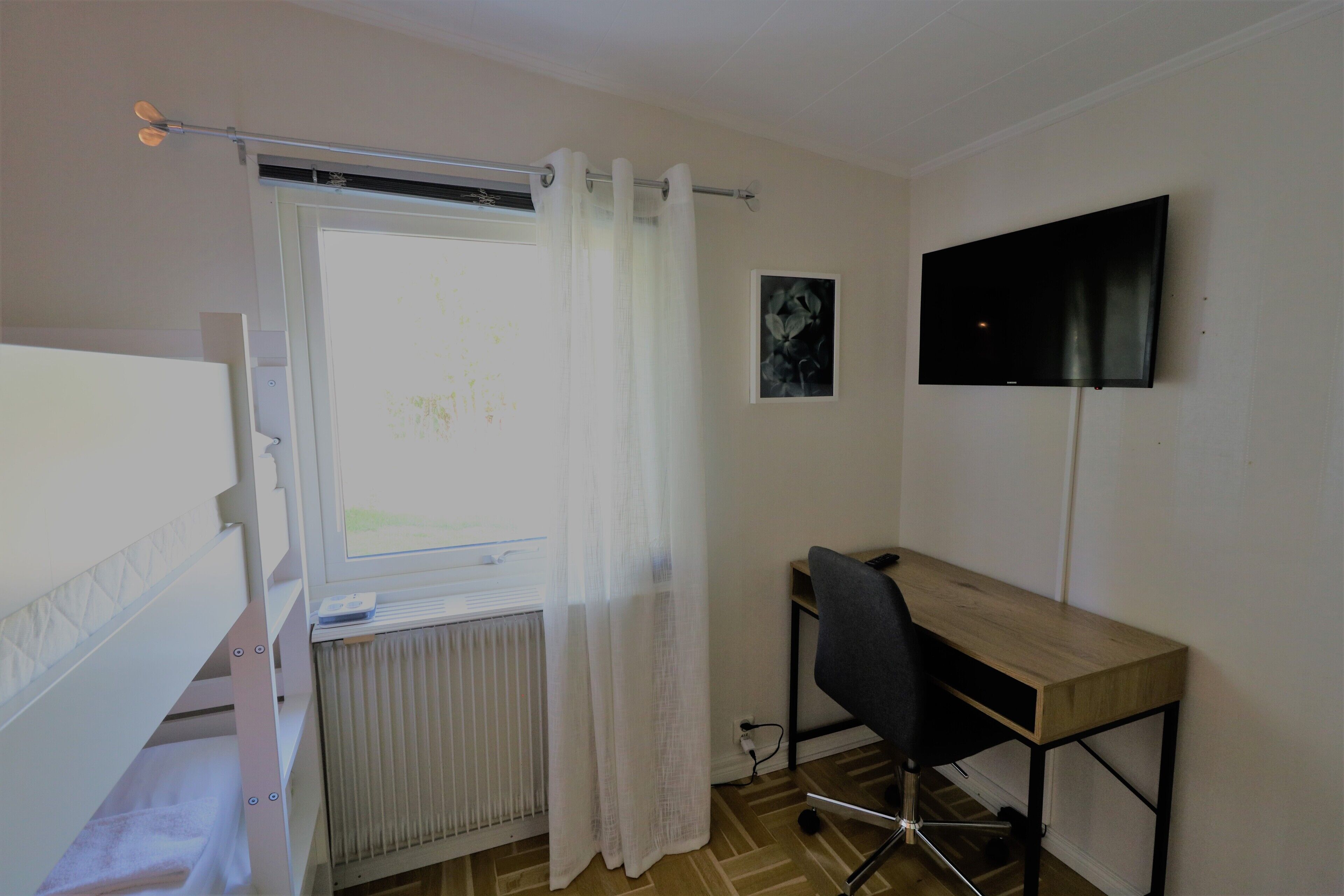 Une chambre avec un lit superposé, un bureau, une chaise, un radiateur et un téléviseur fixé au mur.