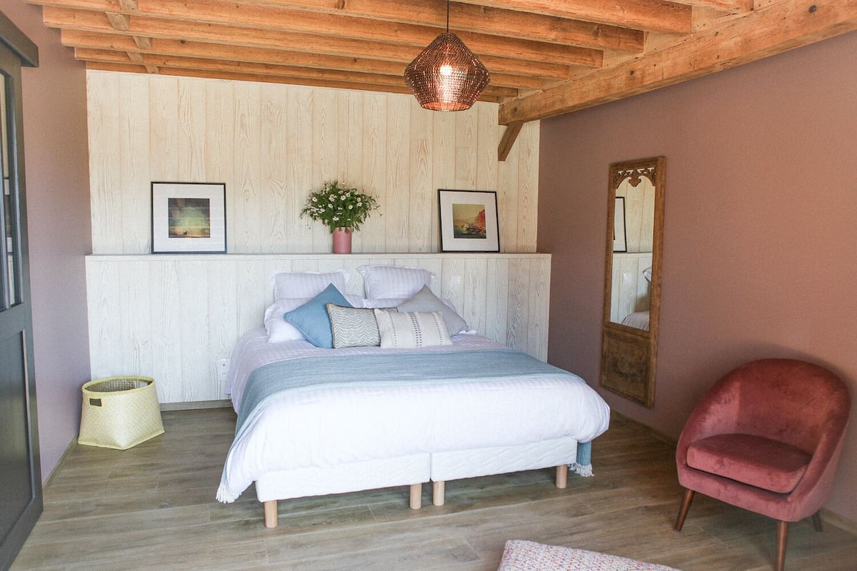Ein Schlafzimmer mit Holzbalkendecke, einem Bett mit weißer Bettwäsche, einem rosa Sessel und einem Spiegel an der Wand.
