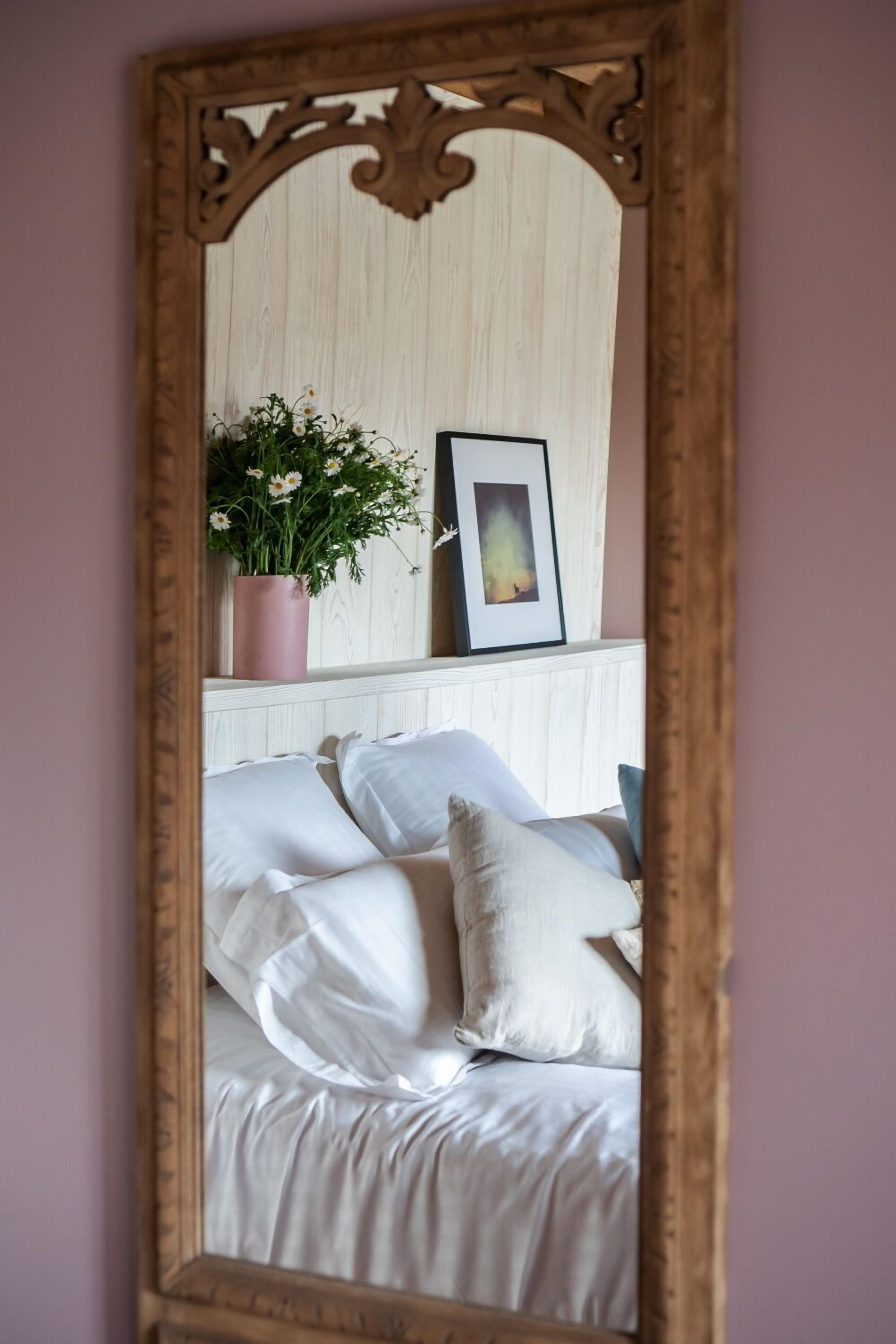 Un dormitorio con cama, almohadas, cómoda, espejo, planta y un cuadro enmarcado.