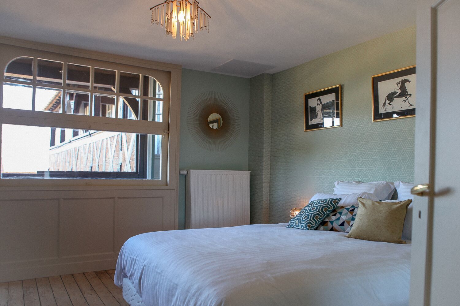 A bedroom with a large window, a bed with patterned pillows, and two framed pictures on the wall.