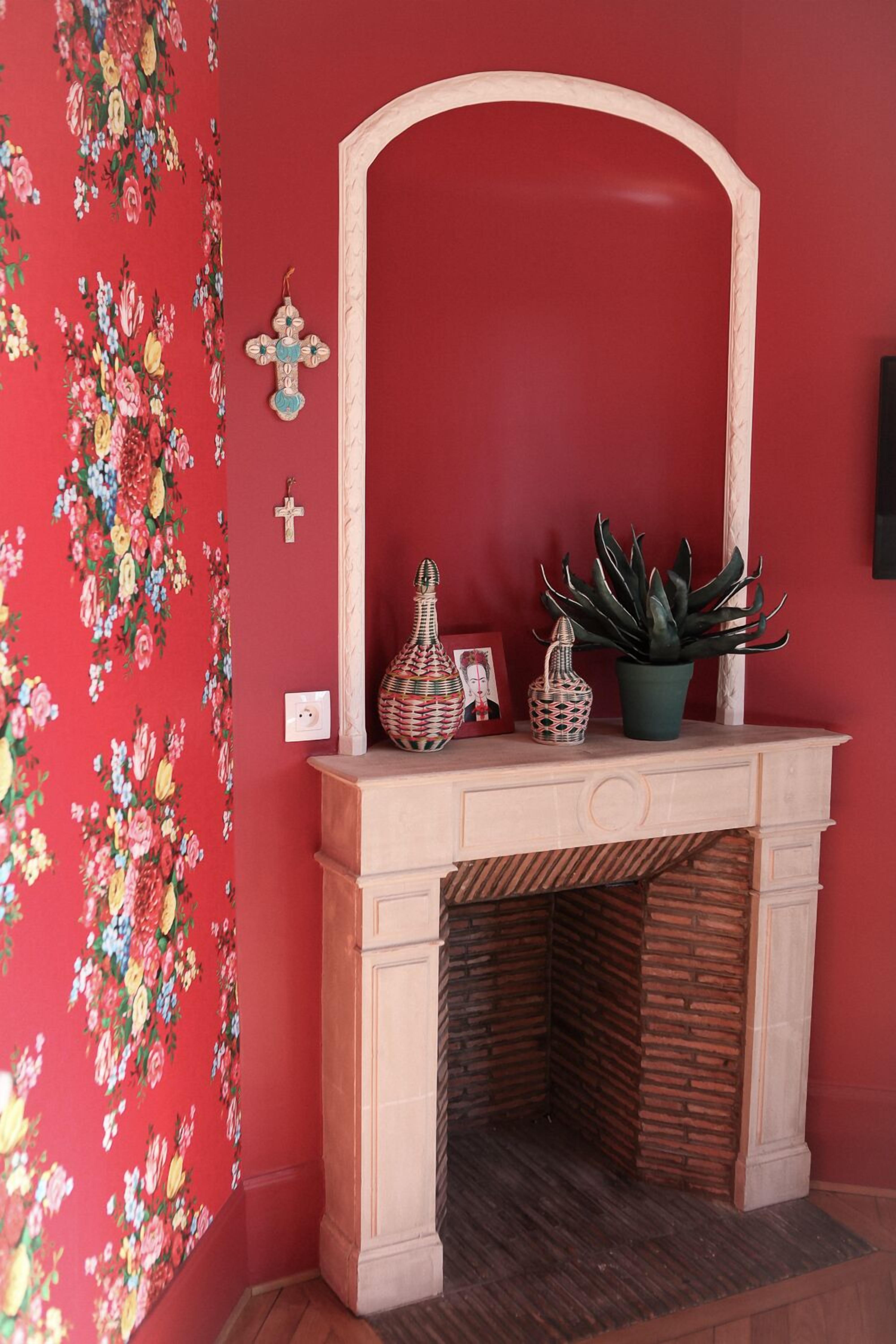 A room with a red floral wallpaper, a wooden mantelpiece with a mirror, and a potted plant.