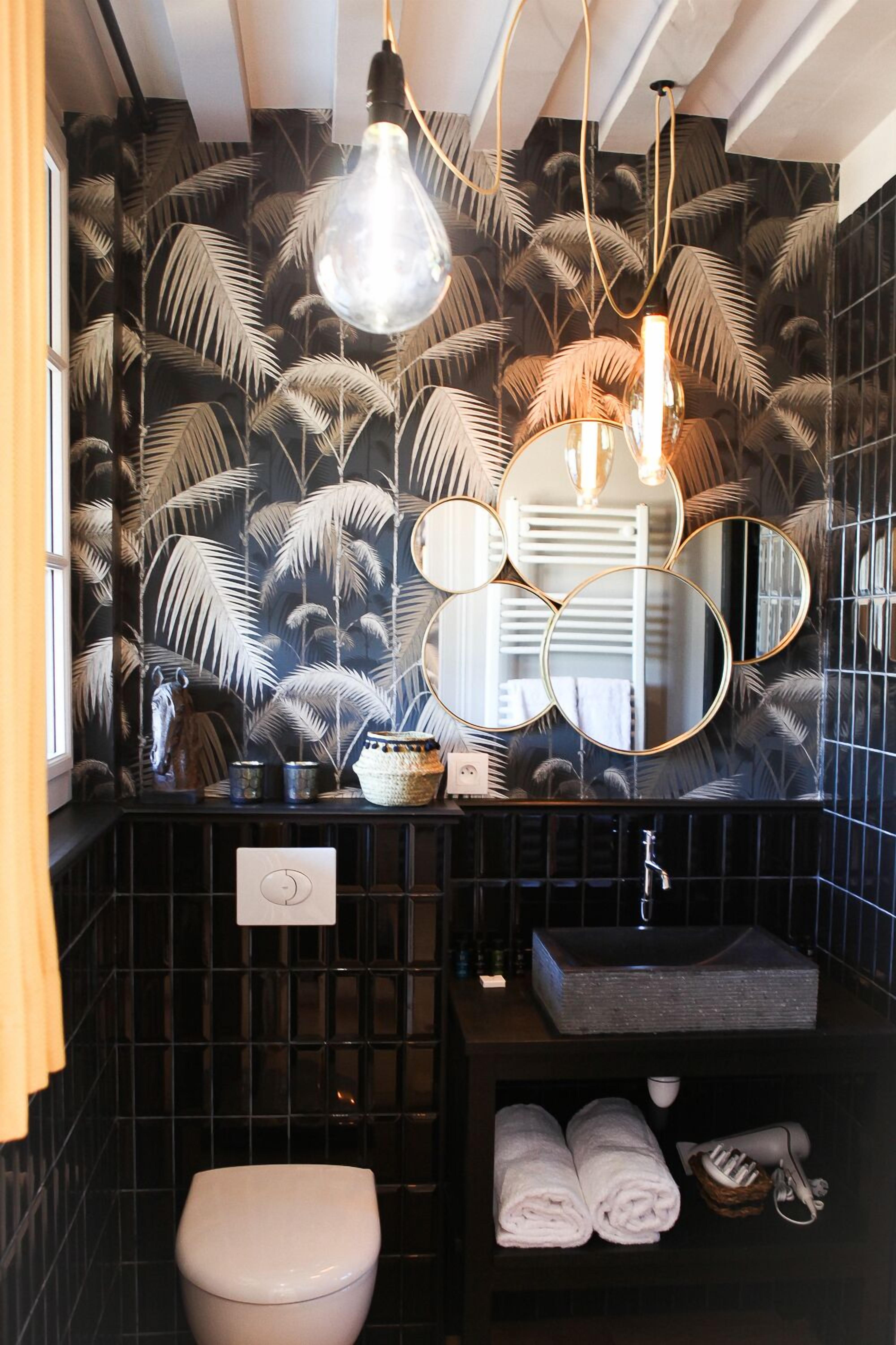 Une salle de bain avec un mur recouvert de carreaux noirs, des toilettes blanches, un lavabo avec un miroir et un porte-serviettes.