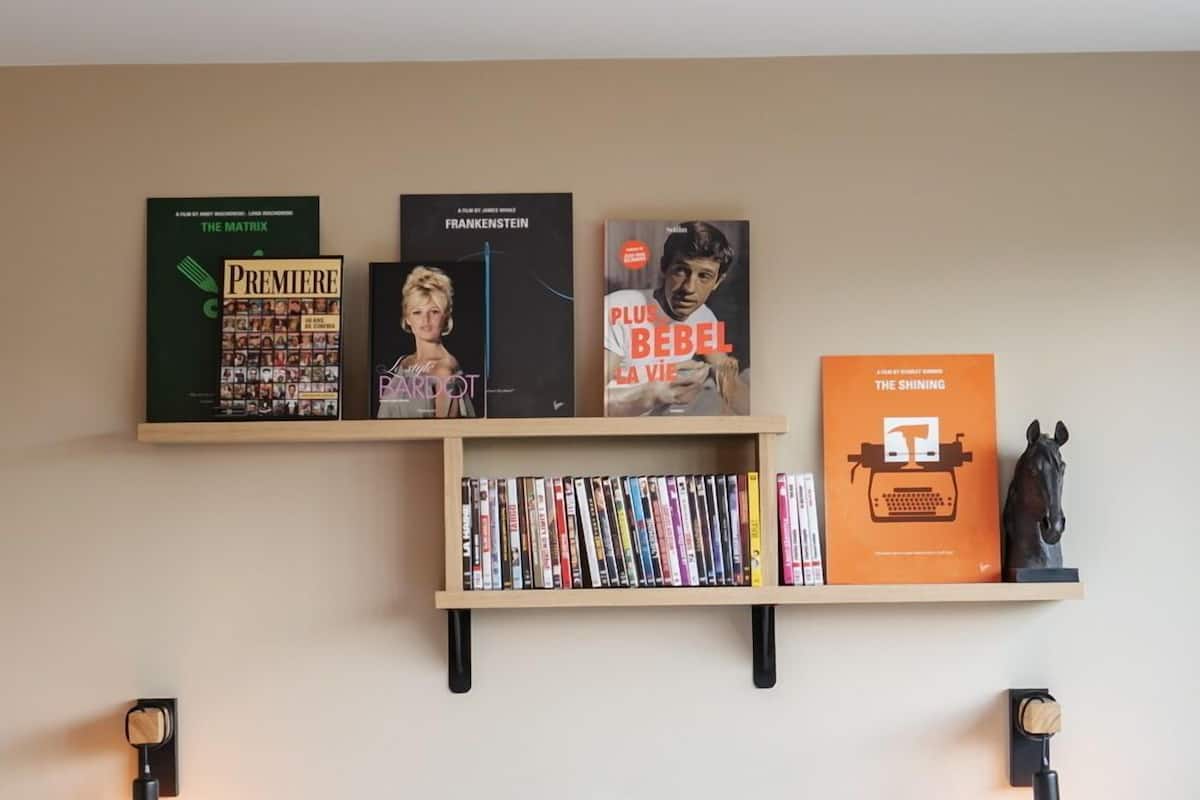 Ein ordentlich eingerichtetes Schlafzimmer mit einem Bett, Kissen, einem Bücherregal mit Büchern und einer Schreibmaschine sowie Wandregalen mit gerahmten Gegenständen.