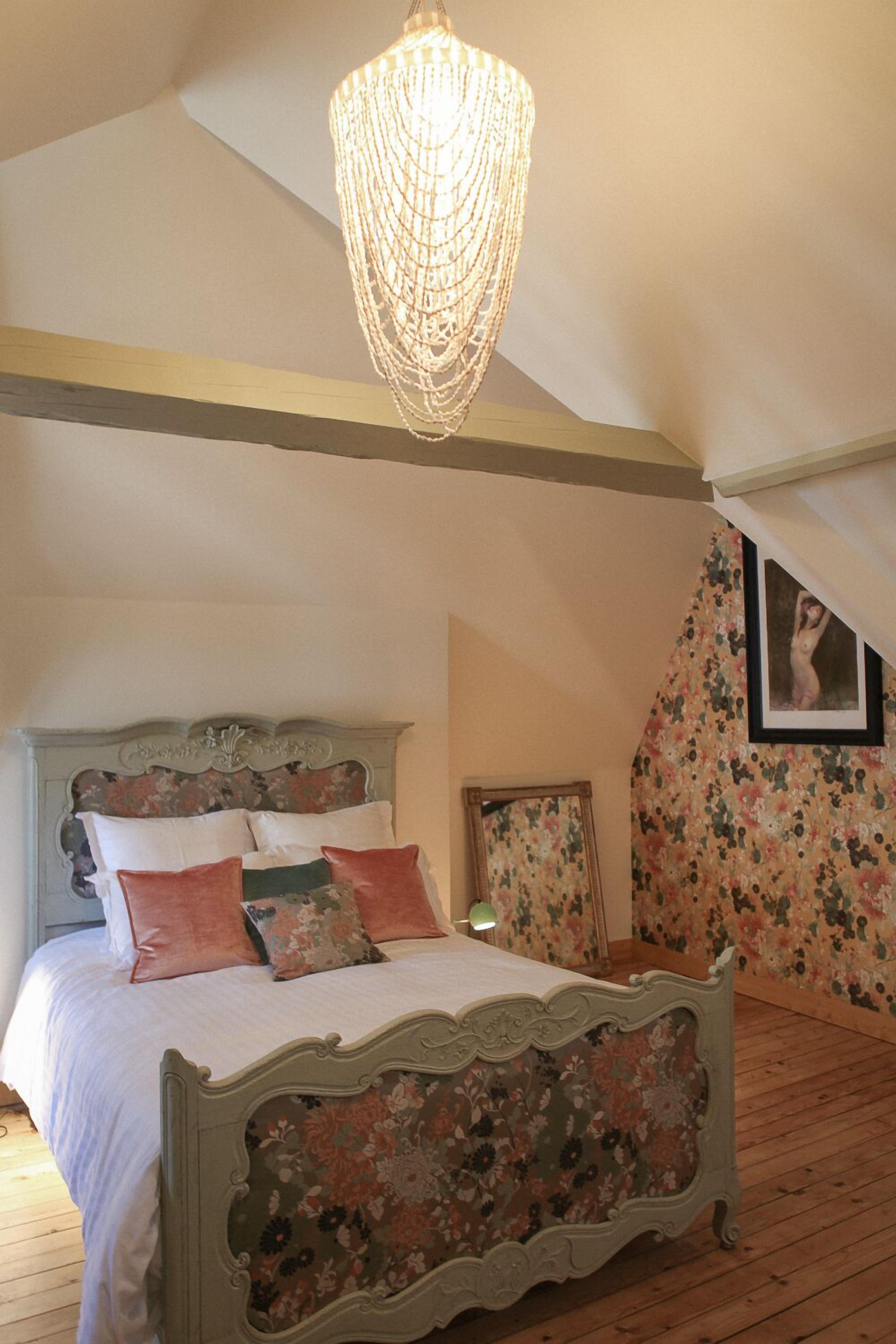 Un dormitorio con cama, papel tapiz floral, una araña y un cuadro en la pared.