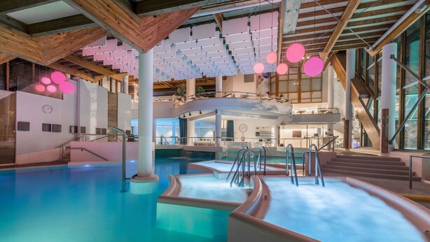Une piscine intérieure à plusieurs niveaux, dotée d’une architecture moderne, avec notamment des poutres en bois et de grandes fenêtres.