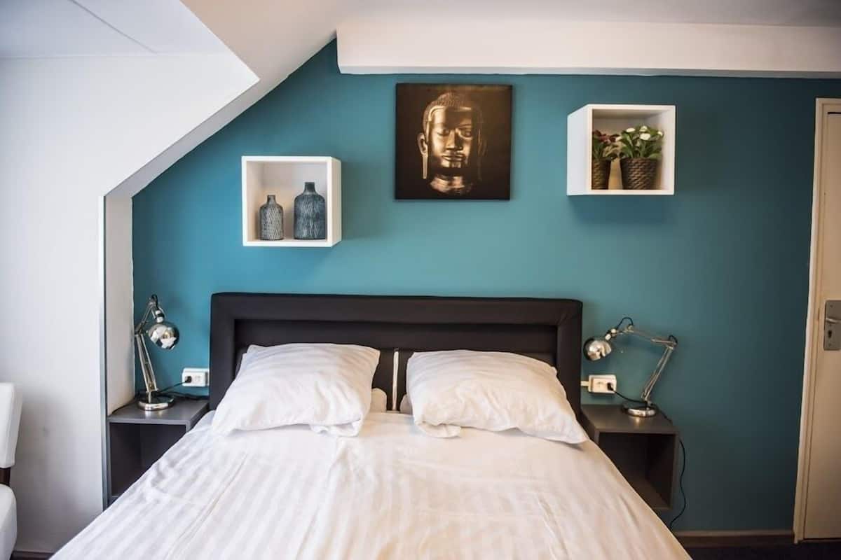 Un dormitorio moderno con cabecera oscura, una cama grande con sábanas blancas, dos mesitas de noche con lámparas, un estante de pared con objetos decorativos y una obra de arte enmarcada sobre la cama.