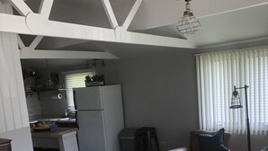 Una sala de estar con un sofá, una mesa de centro, un tapete estampado y una zona de cocina con un refrigerador y taburetes de bar.