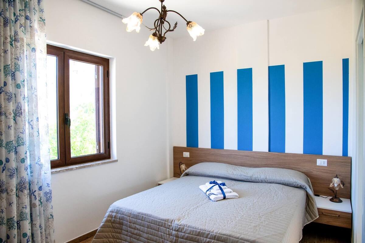 Ein Schlafzimmer mit Bett, Nachttisch, Wandlampe und einem Fenster mit Vorhängen.