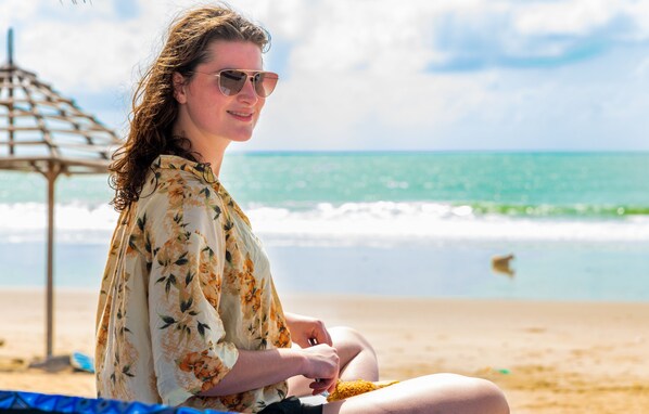 Uma pessoa sentada na praia, com uma estrutura de madeira ao fundo.