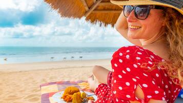 Una persona sentada en la playa con un plato de comida, usando un vestido de lunares y un sombrero de paja.