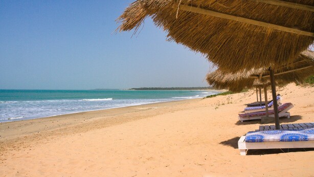 Una playa con una sombrilla de paja y sillones de descanso.