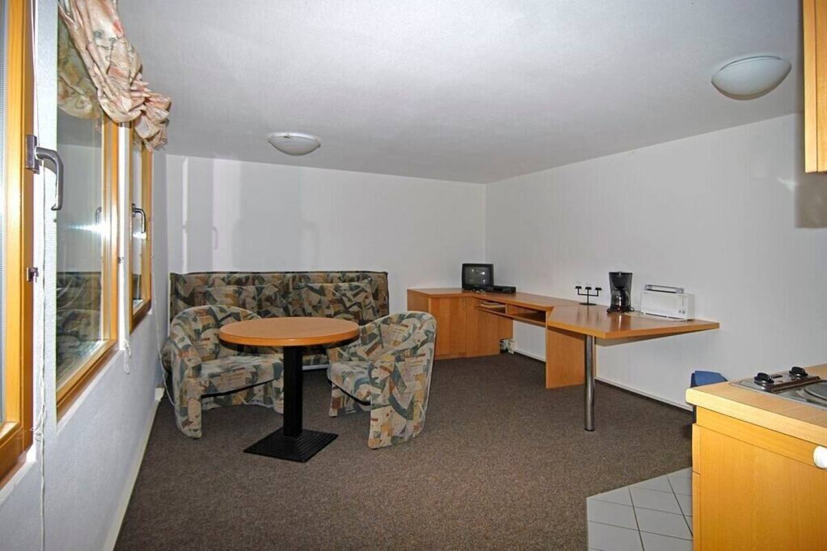A compact room with a sofa, a small round table, a desk with a computer, and a kitchenette area.