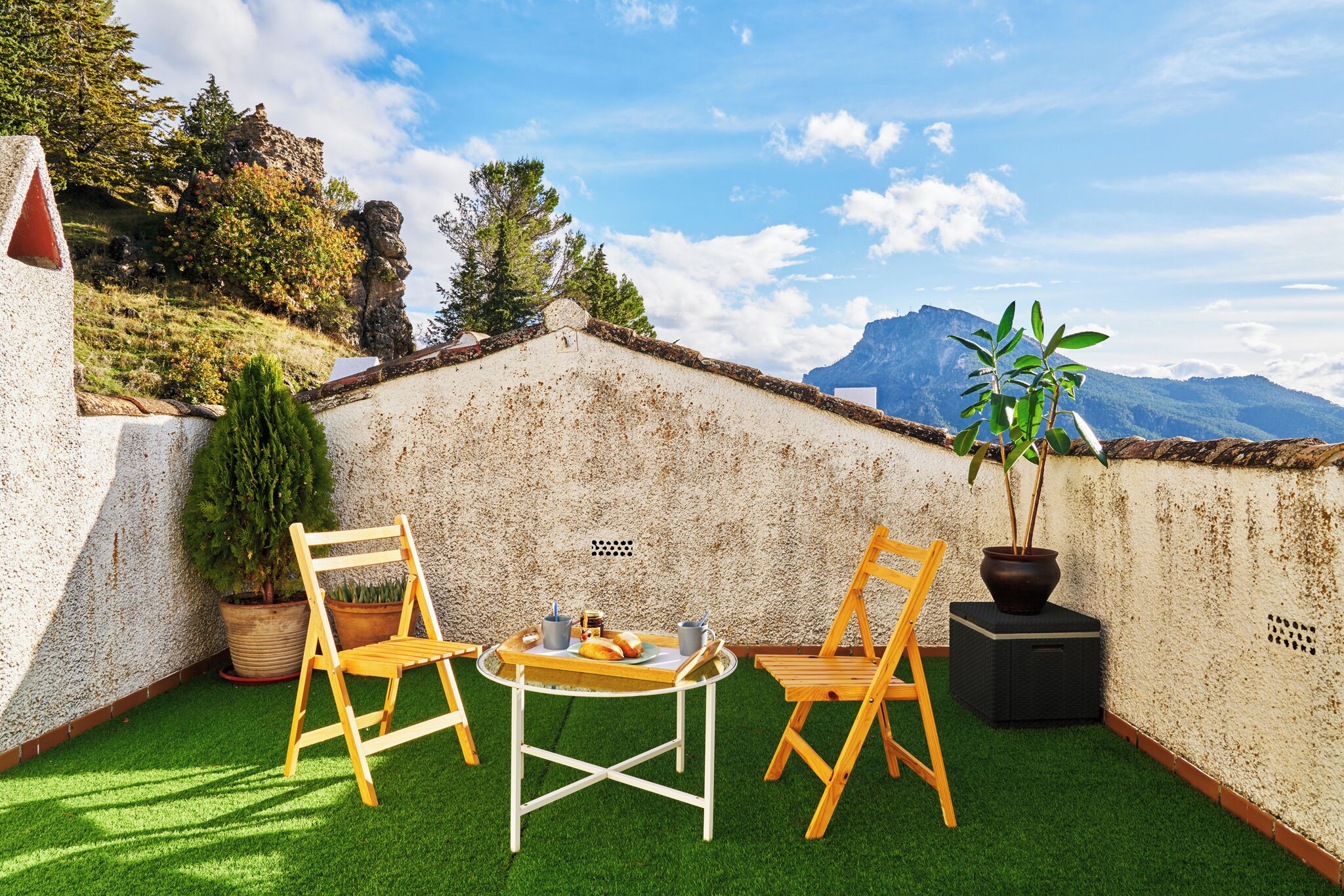 Een terras op het dak, met een tafel, twee stoelen, een plant in pot en uitzicht op bergen.