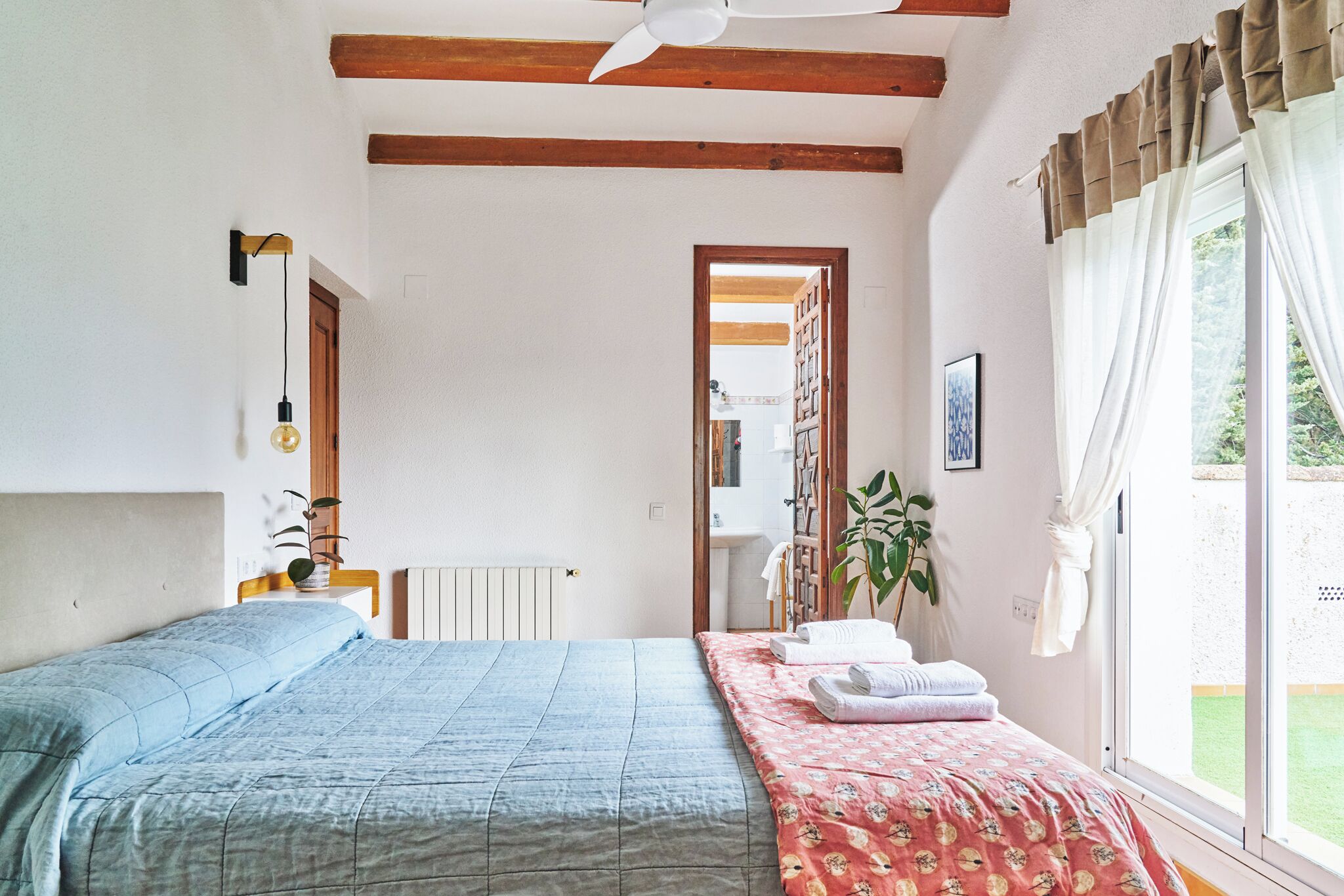 Ein Schlafzimmer mit einem Bett, einem Heizkörper, einem Fenster mit Vorhängen, einer Pflanze und einer Tür zu einem Badezimmer.