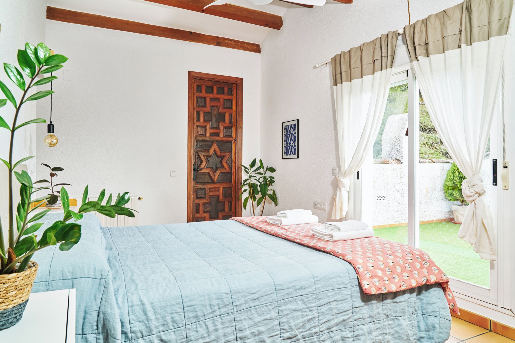 A bedroom with a bed, a wooden door, a potted plant, and a view of a garden through the window.
