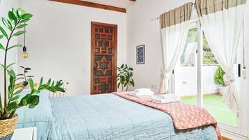 A bedroom with a bed, a wooden door, a potted plant, and a view of a garden through the window.