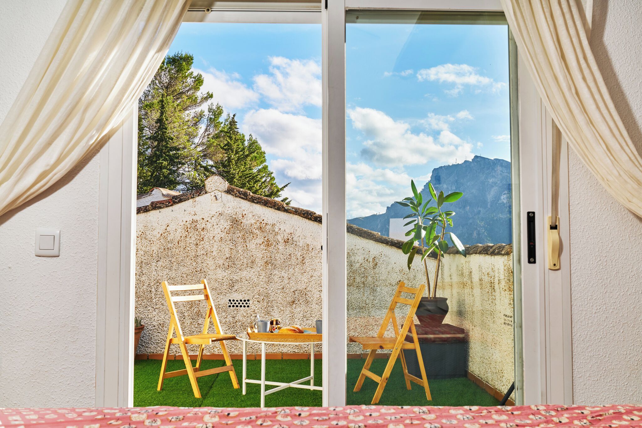 A room with a sliding glass door leading to an outdoor area with two wooden chairs, a small table, and a potted plant.