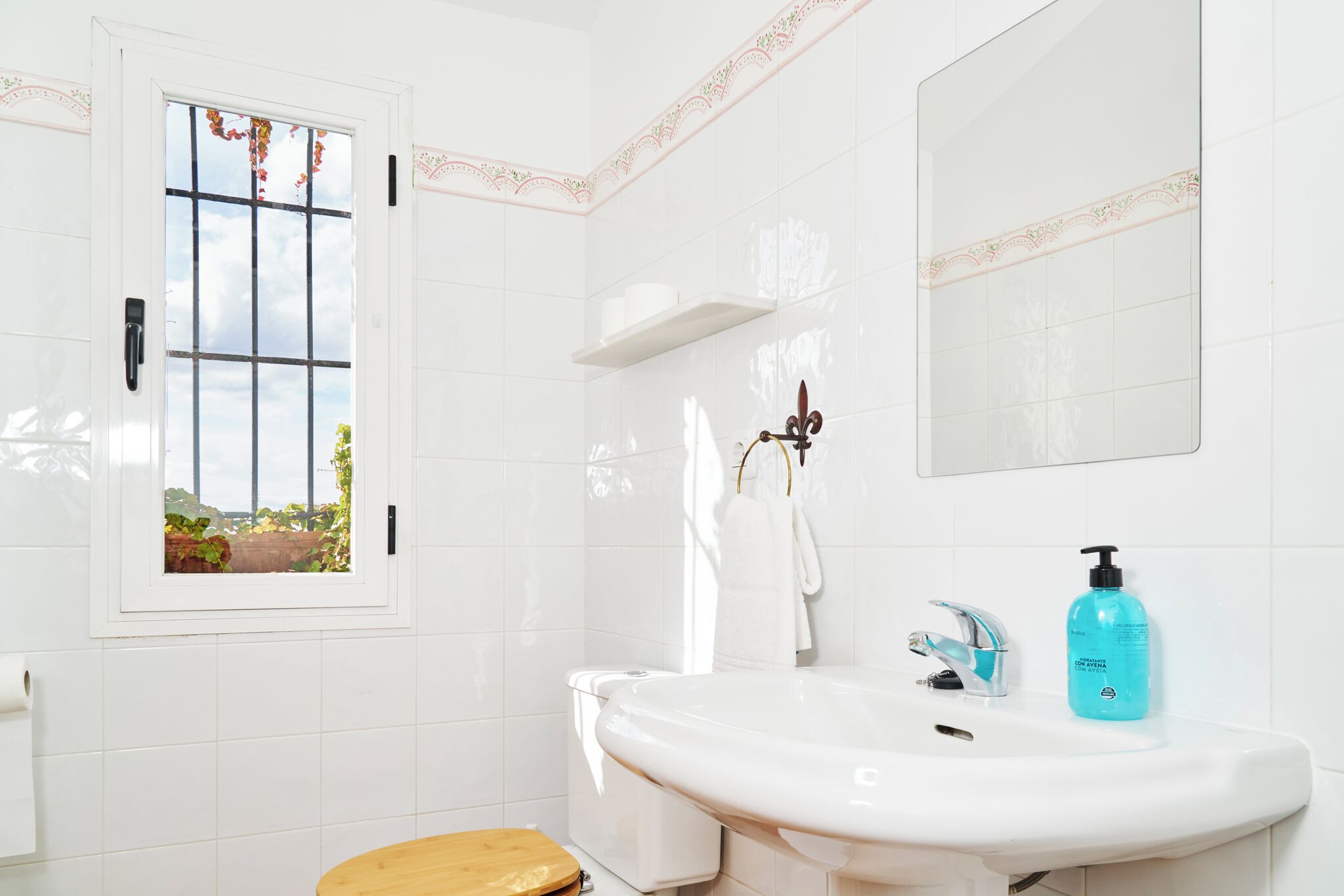 Une salle de bain avec un lavabo blanc, un miroir, une fenêtre donnant sur un espace verdoyant et des toilettes.