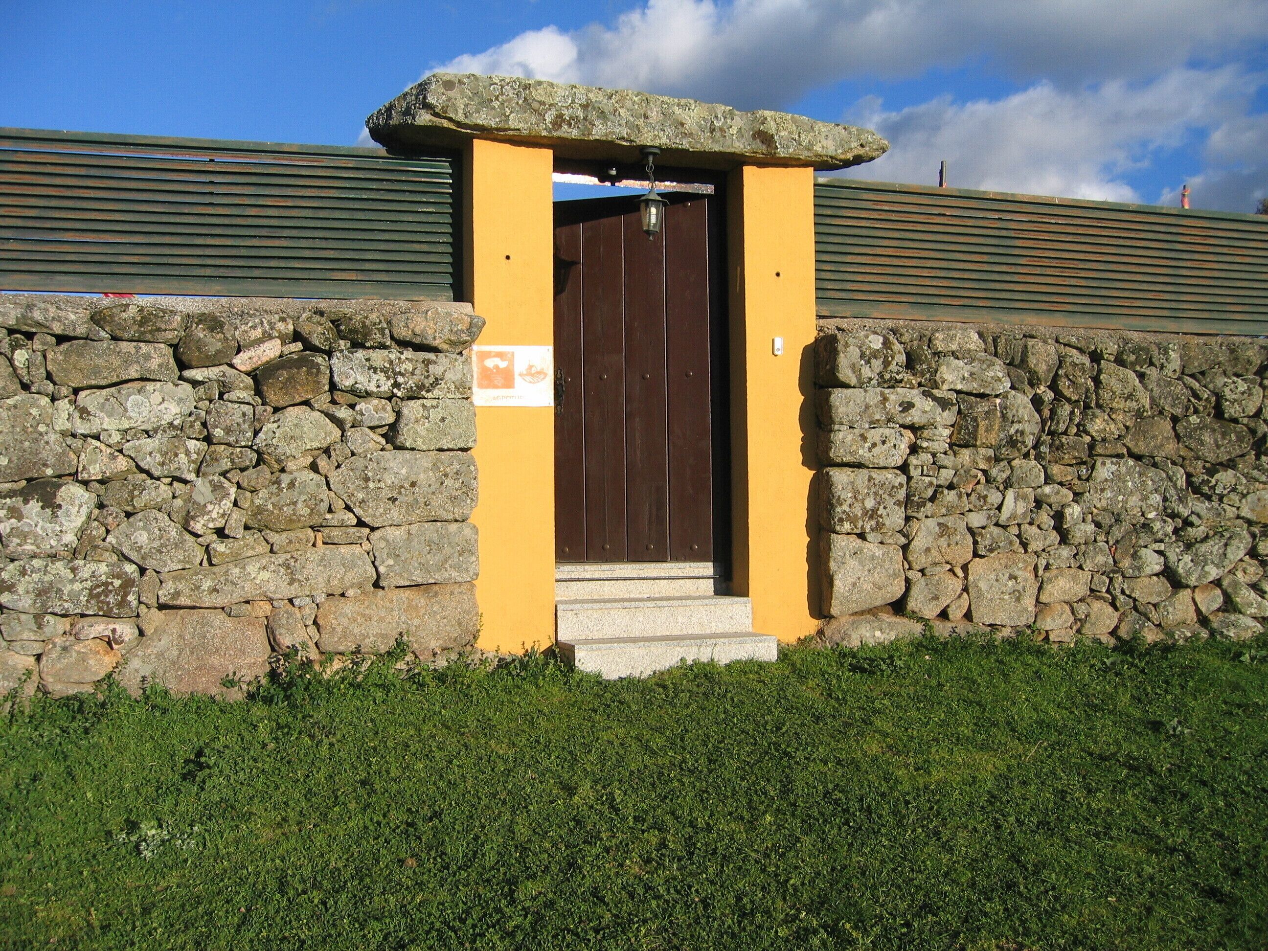 Una entrada de pared de piedra con una puerta de madera, una pequeña estructura de piedra encima de la puerta y un área cubierta de césped al frente.