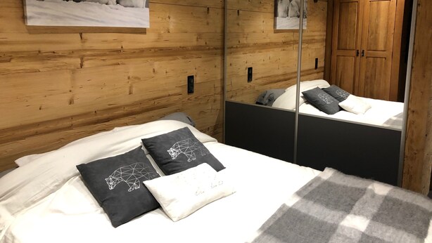A wooden-paneled room with a bed, pillows, a mirror, and a wall-mounted lamp. - BEAUTIFUL T3 ESPRIT CHALET (LES ANGLES)