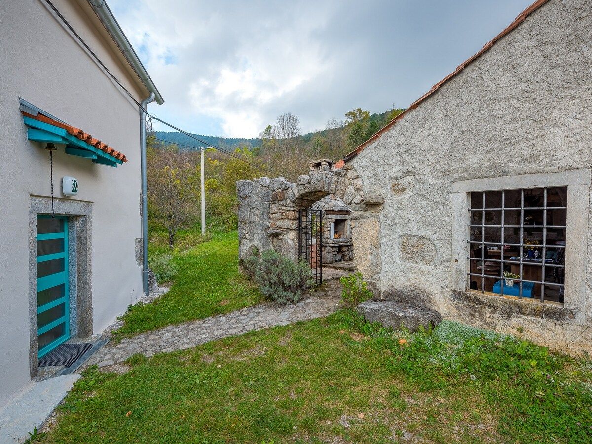 Un étroit sentier de pierres mène à un bâtiment rustique en pierre, doté d’un portail en métal et d’une fenêtre grillagée.