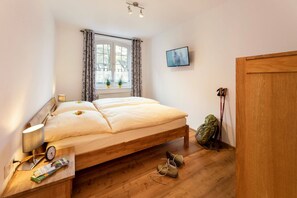 A bedroom with a wooden bed, a window with curtains, a TV on the wall, and a pair of shoes and a backpack on the floor.