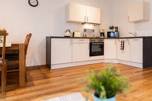 A modern kitchen with white cabinets, a built-in oven, and a microwave. There is a dining table with chairs and a clock on the wall.