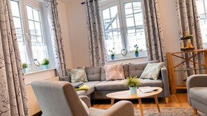 A modern living room with a grey sofa, two armchairs, a round wooden coffee table, and a small side table. There are curtains with a leaf pattern, a window with a view of buildings, and a shelf with plants and a decorative item. - Vacation apartment "Katarina 1", right next to Landhotel Albers - your landlords  (Schmallenberg)