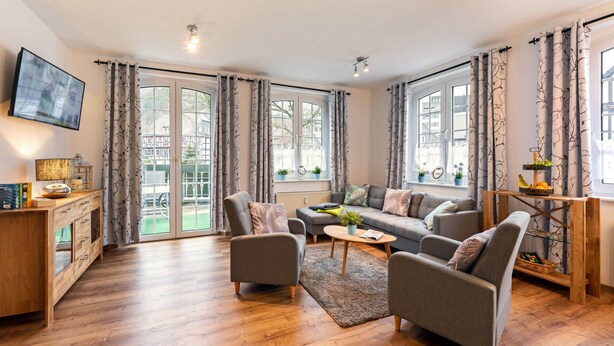 A modern living room with a sofa, armchairs, a coffee table, and a TV. There are large windows with curtains, a wooden floor, and a view of the outside. - Vacation apartment "Katarina 1", right next to Landhotel Albers - your landlords  (Schmallenberg)
